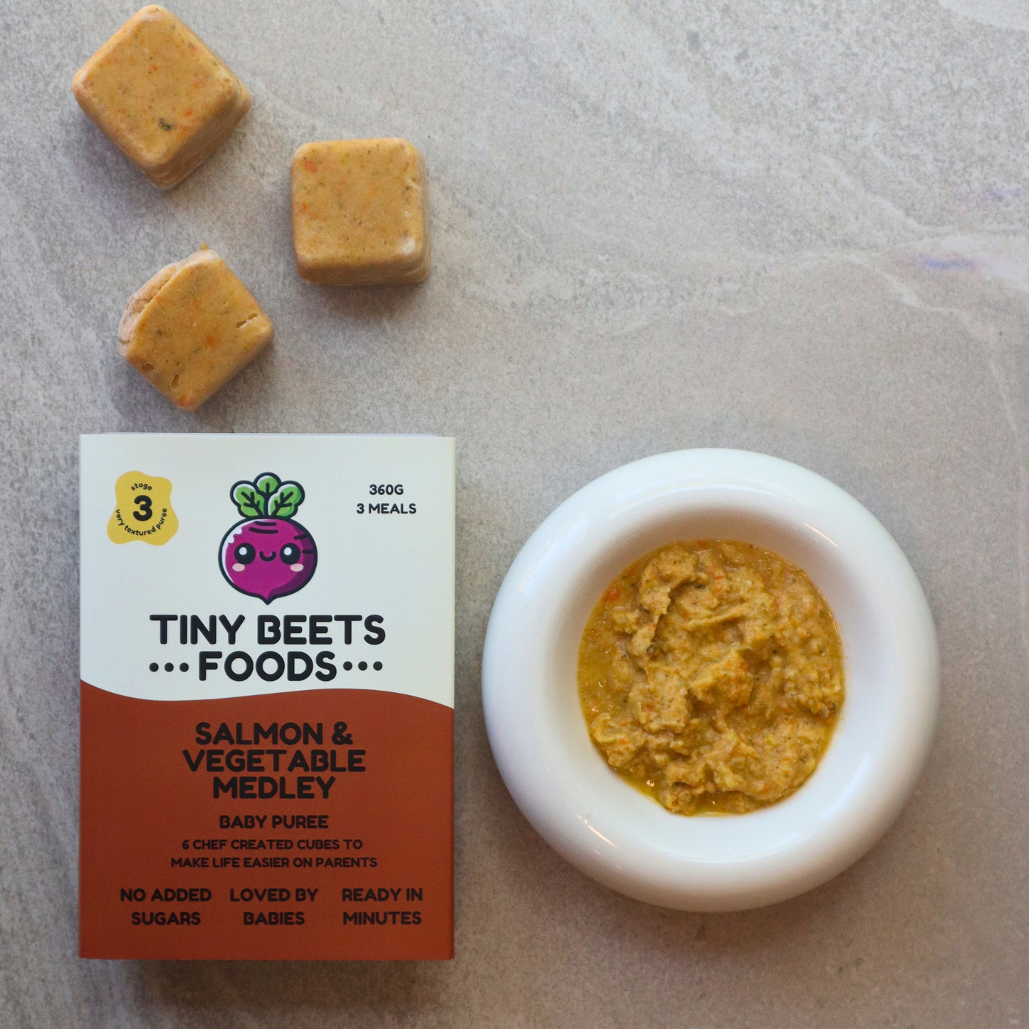 Tiny Beets Foods salmon and vegetable medley puree with packaging and a small bowl of puree on a neutral background.