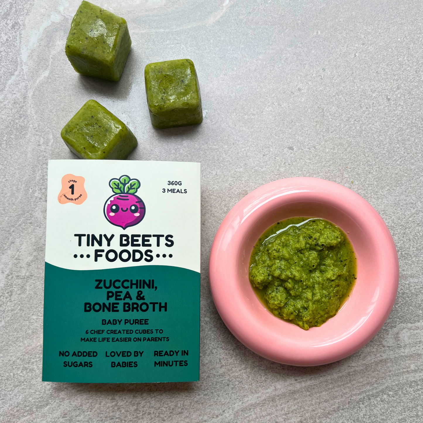Zucchini, pea, and bone broth baby puree cubes with packaging and a pink bowl of puree on a gray surface.