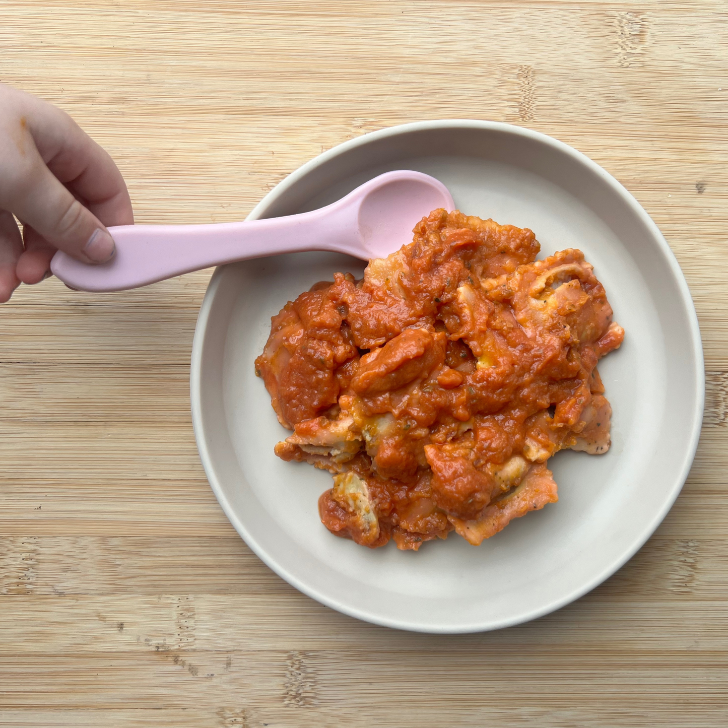 
                  
                    A hand holding a pink spoon next to a plate of chicken ravioli in a red sauce, set on a wooden surface.
                  
                