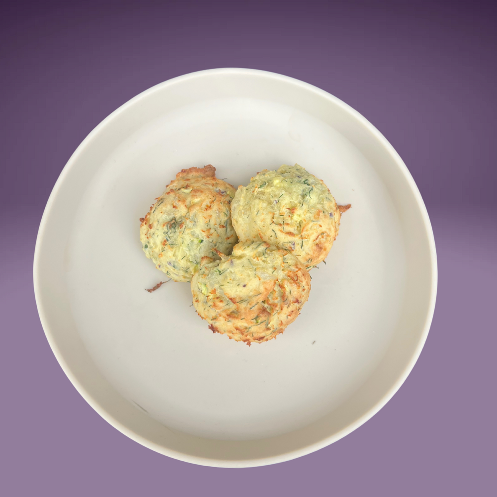 Three golden-baked Fish, Potato & Dill Balls served in a white bowl, set against a soft purple background. The balls have a light, fluffy texture with visible herbs and finely grated vegetables, ideal for baby-led weaning.